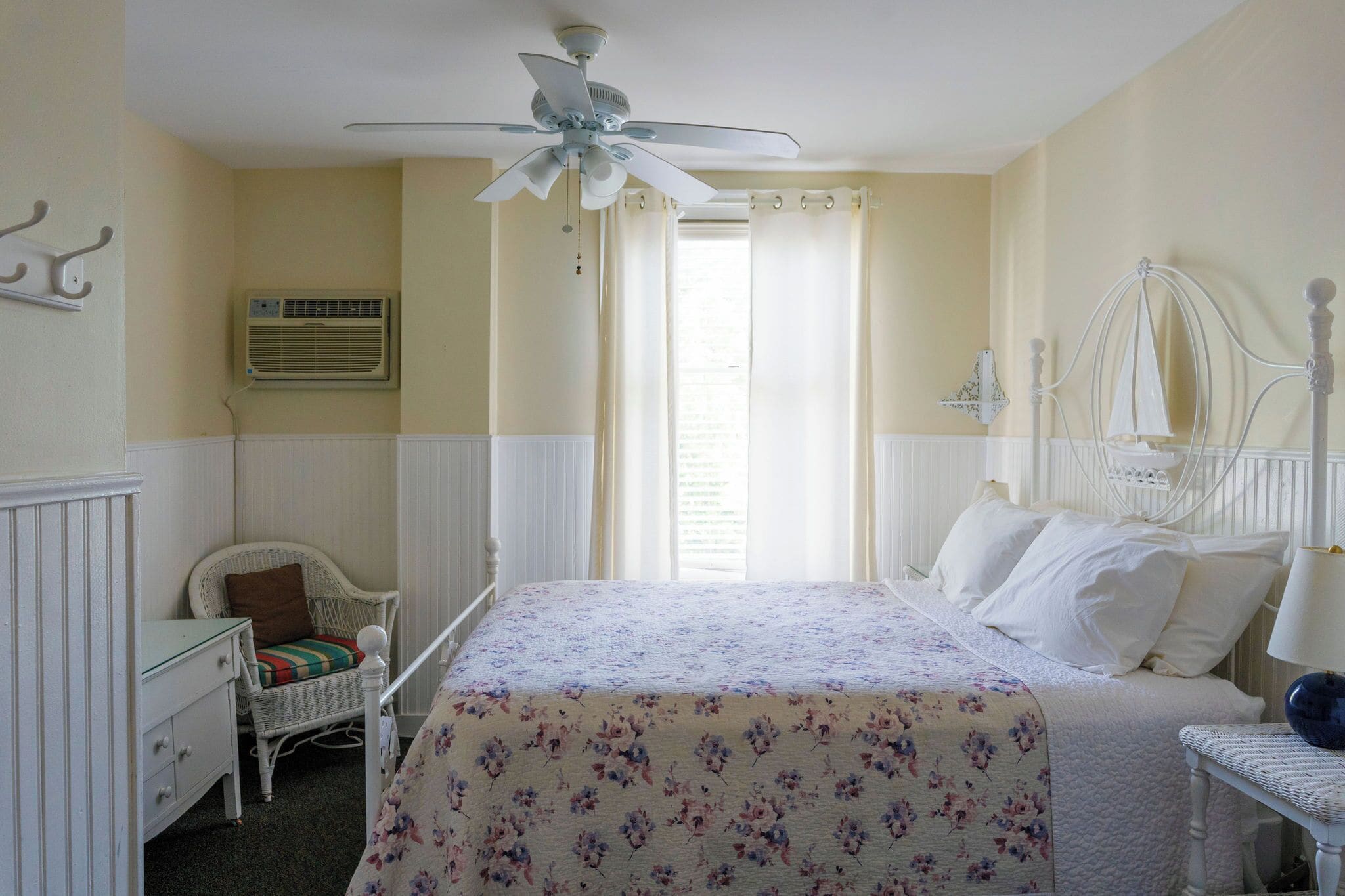 A bedroom with a bed, a chair, a nightstand, a ceiling fan, and a window with curtains.