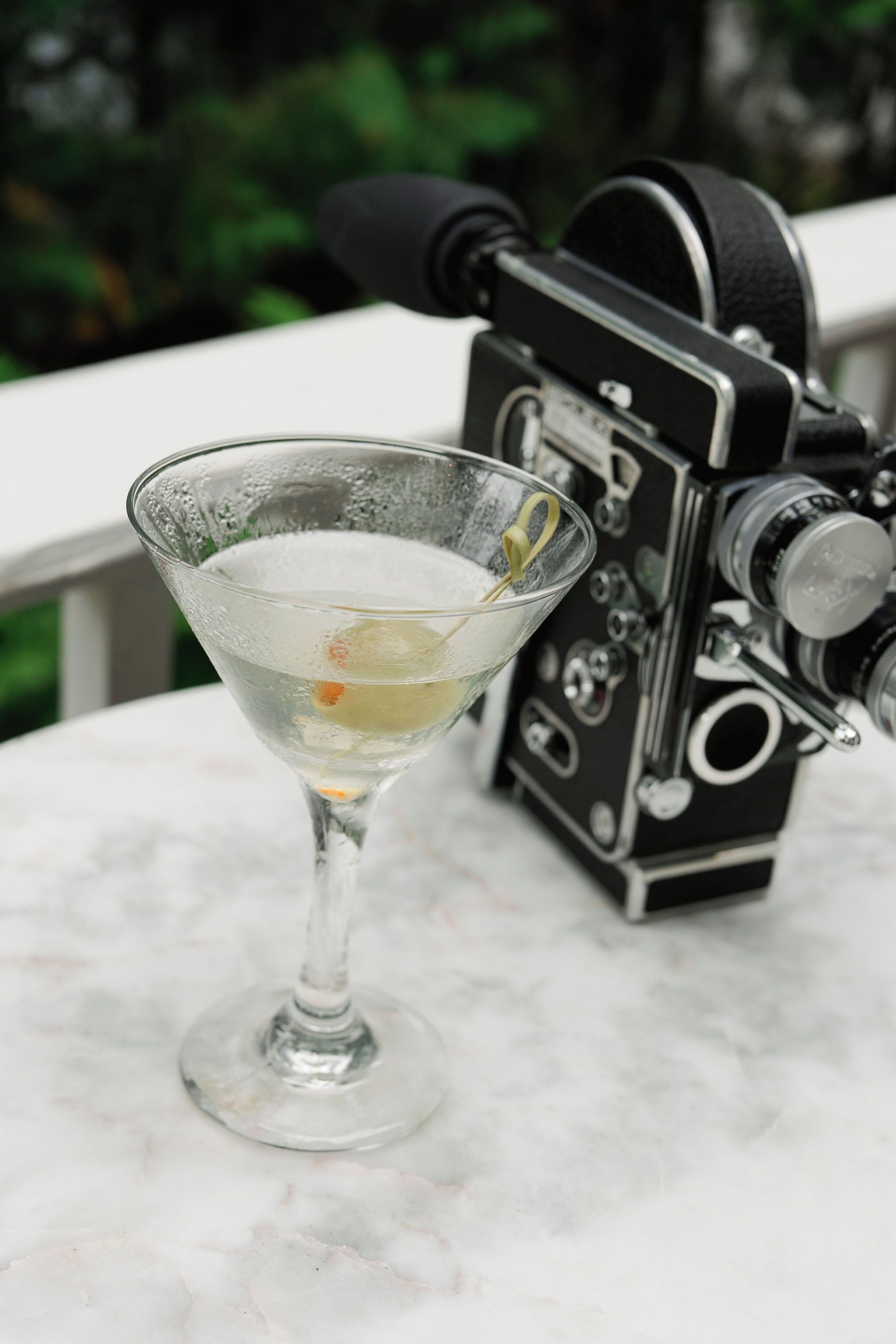 A martini glass with a lemon twist on a marble table. A vintage camera is positioned behind the glass.