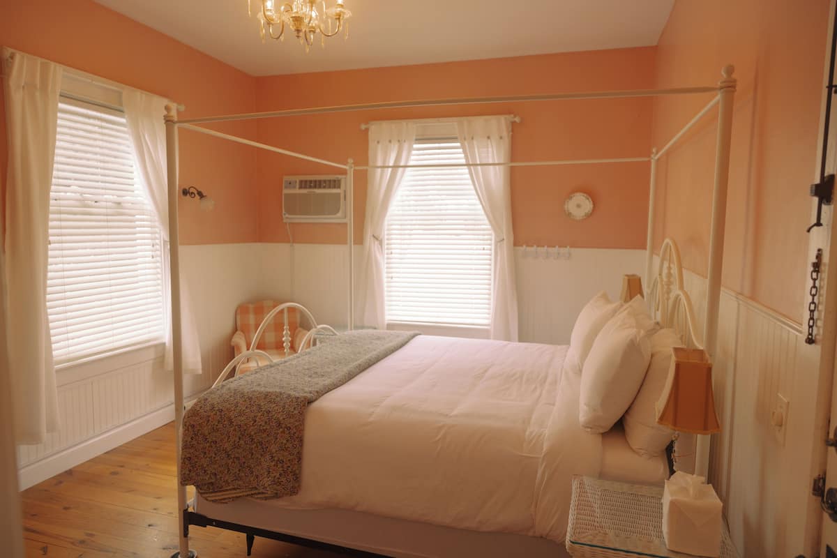 A bedroom with a four-poster bed, a bedside table with a lamp, and a window with blinds.