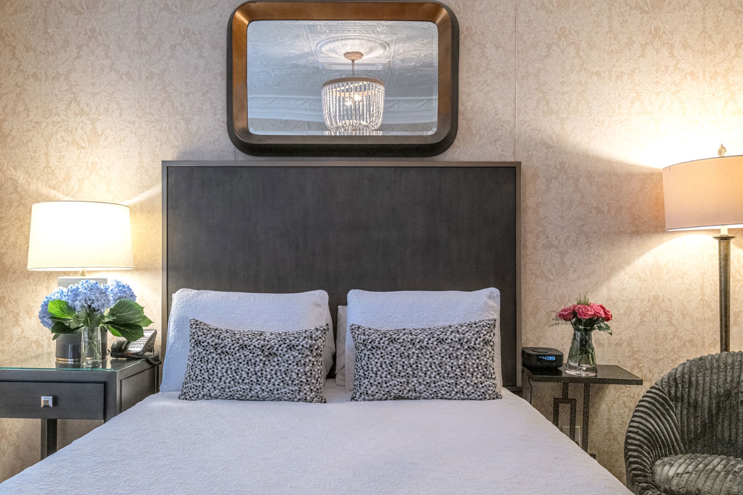 Habitación de hotel con una cama grande, dos almohadas, una mesita de noche con una lámpara, un jarrón con flores e imagen enmarcada sobre la cama.