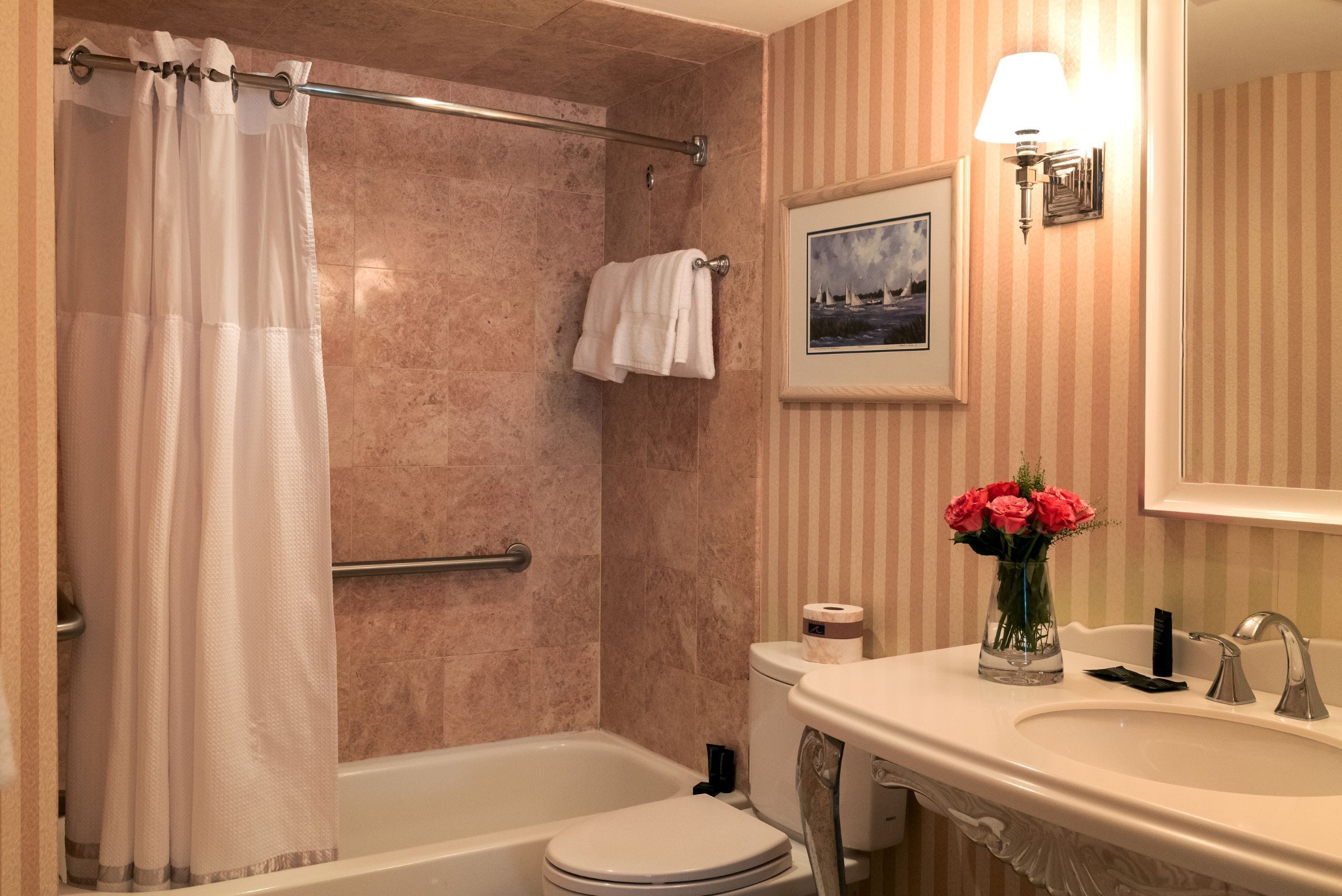 Une salle de bain avec une baignoire, des toilettes et un lavabo. On y trouve un rideau de douche, un porte-serviettes, un tableau encadré au mur et un vase avec des fleurs sur le lavabo.