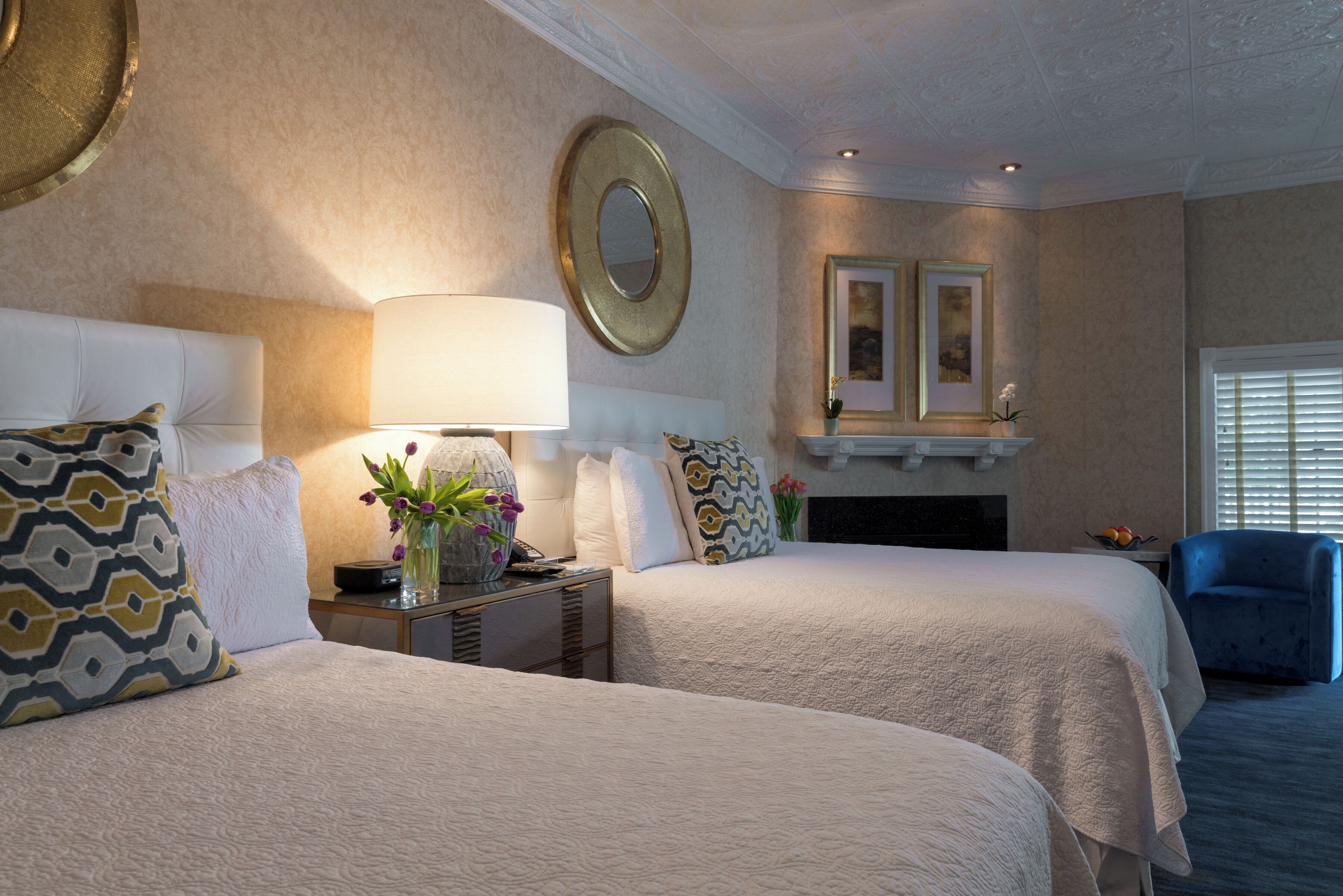 A hotel room with two beds, a nightstand, a lamp, a mirror, and a fireplace.