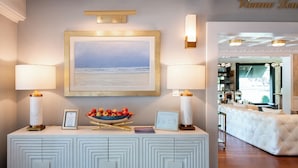 Une pièce avec un canapé blanc à motifs capitonnés, un tableau encadré représentant une plage, et une console blanche sur laquelle sont posés un saladier de fruits et un certificat encadré.