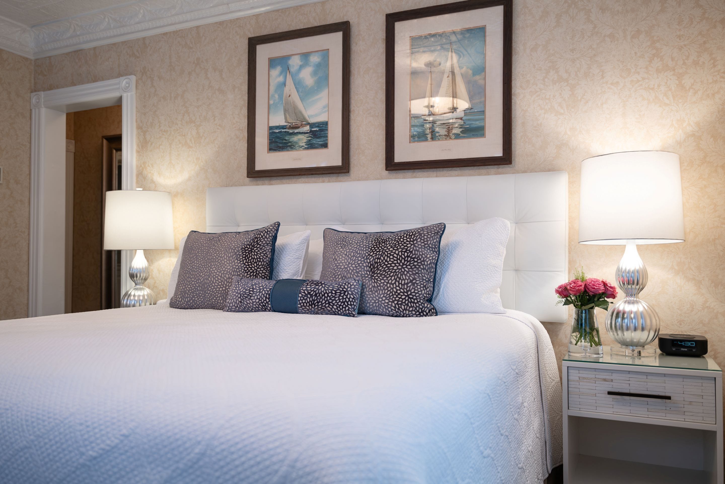 A hotel room with a large bed, two framed paintings, a nightstand with a lamp, and a vase with flowers.