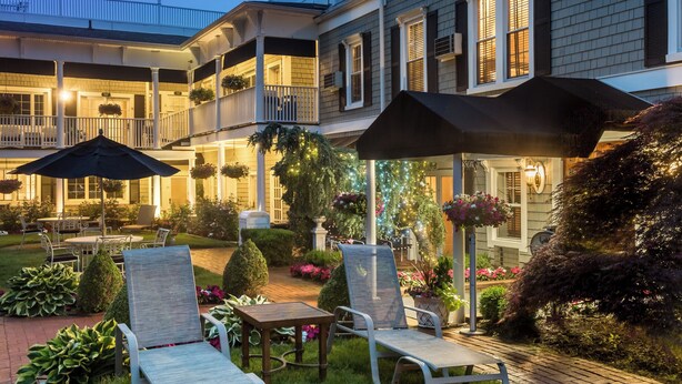 Un bâtiment à deux étages doté d’un balcon, d’un espace extérieur aménagé avec des chaises et des tables, et d’un jardin bien entretenu où poussent diverses plantes et fleurs.