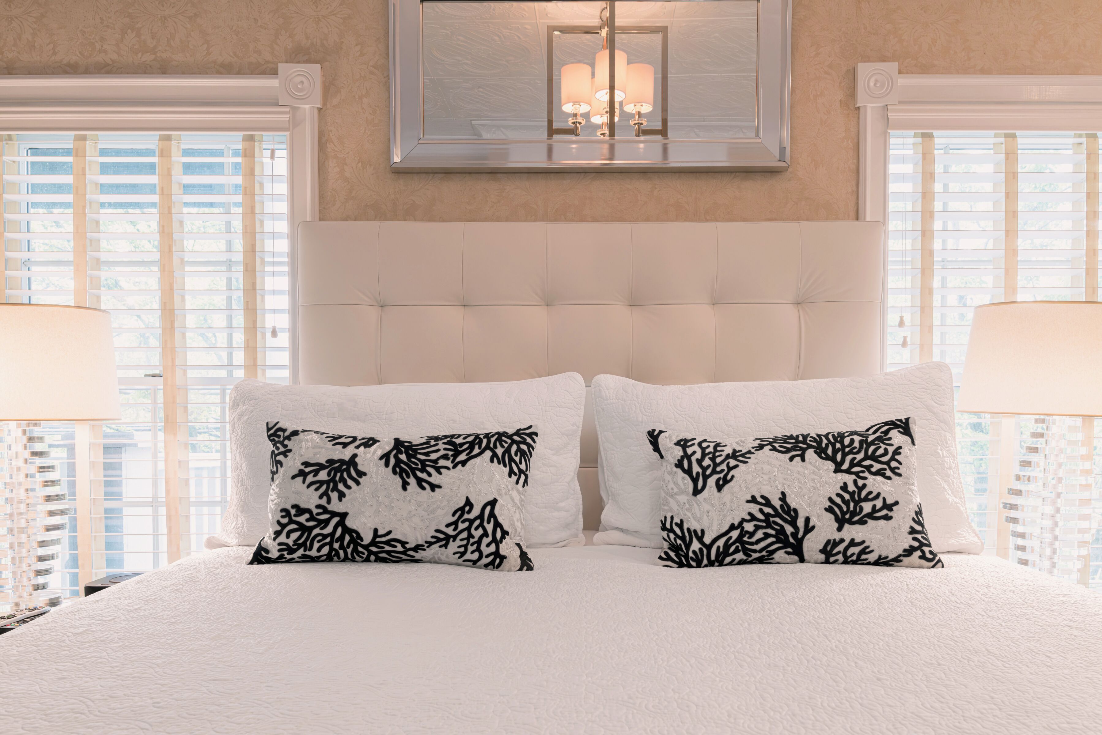 Un dormitorio con cabecera capitonada, dos almohadas blancas con motivos de coral negro y un candelabro colgante.