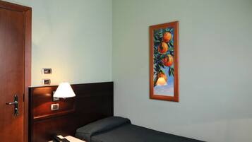 A single bed with a dark bedspread, a bedside table with a lamp, a telephone, and a small drawer unit. A painting of oranges hangs on the wall.