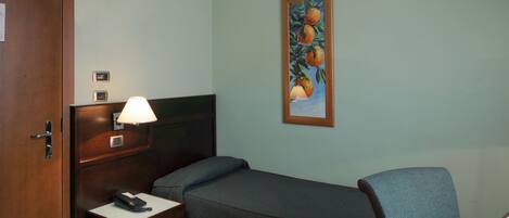 A hotel room with a single bed, bedside table with a lamp, a desk with a chair, and a painting on the wall.