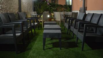 An outdoor seating area with wicker furniture and a small table, surrounded by a garden and a building wall.
