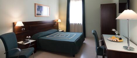 A hotel room with a bed, two bedside tables, a desk with a chair, a lamp, a mirror, and a window with curtains.