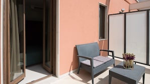 Un balcón con una silla de mimbre, una pequeña mesa, un ventanal corredero y una pared con un ventanal.