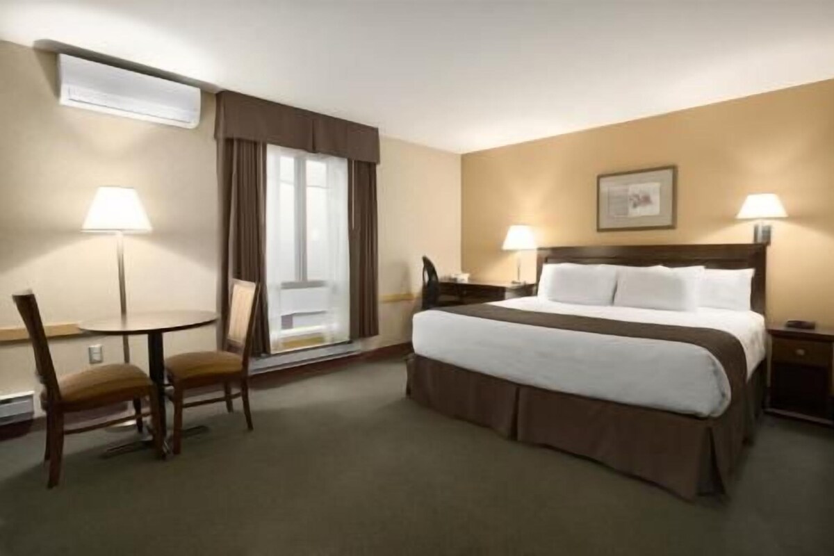Quarto de hotel com cama, escrivaninha, cadeira, janela com cortinas e duas luminárias de cabeceira.