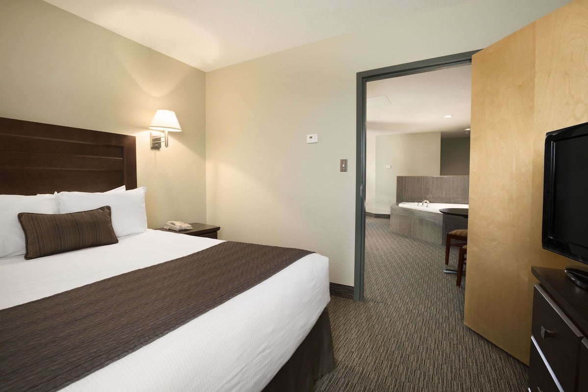 A hotel room with a large bed, a desk with a lamp, a TV, and a bathroom visible through an open door.
