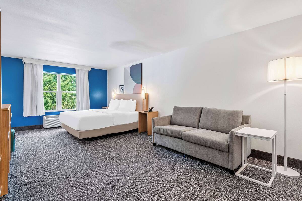 A hotel room with a bed, a sofa, a small table, a lamp, and a window with curtains.