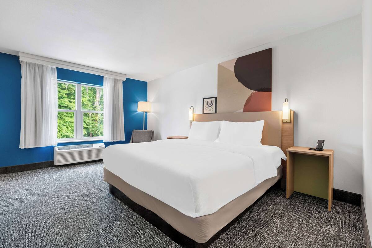 A hotel room with a large bed, a bedside table with a lamp, a wall-mounted artwork, and a window with curtains.