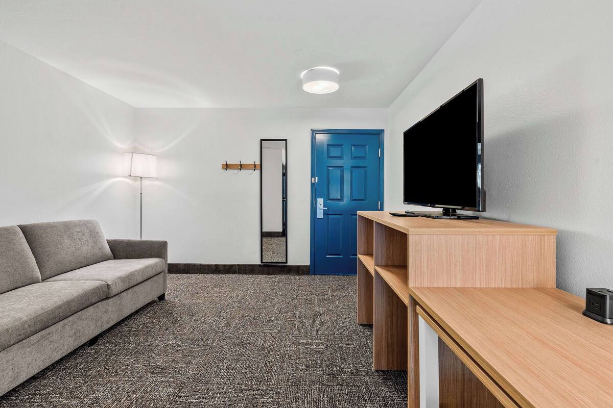 A modern hotel room with a grey sofa, a wooden TV stand, a flat-screen TV, a blue door, and a floor lamp.
