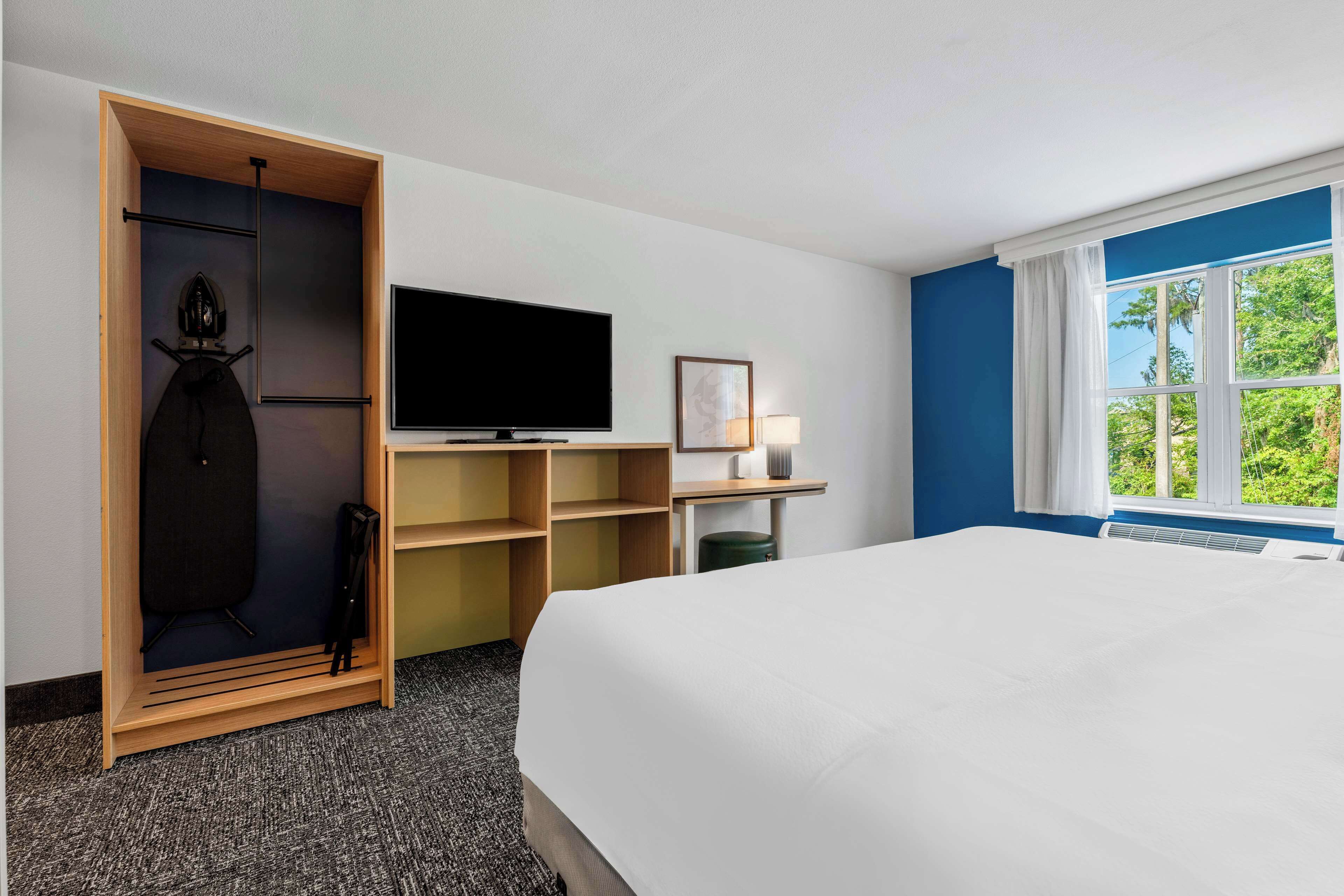 A hotel room with a bed, a television, a desk with a lamp, a wardrobe, and a window with a view of greenery.