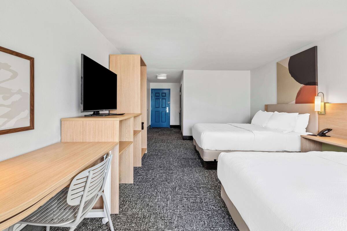 A hotel room with a desk, two beds, a TV, and a blue door in the background.
