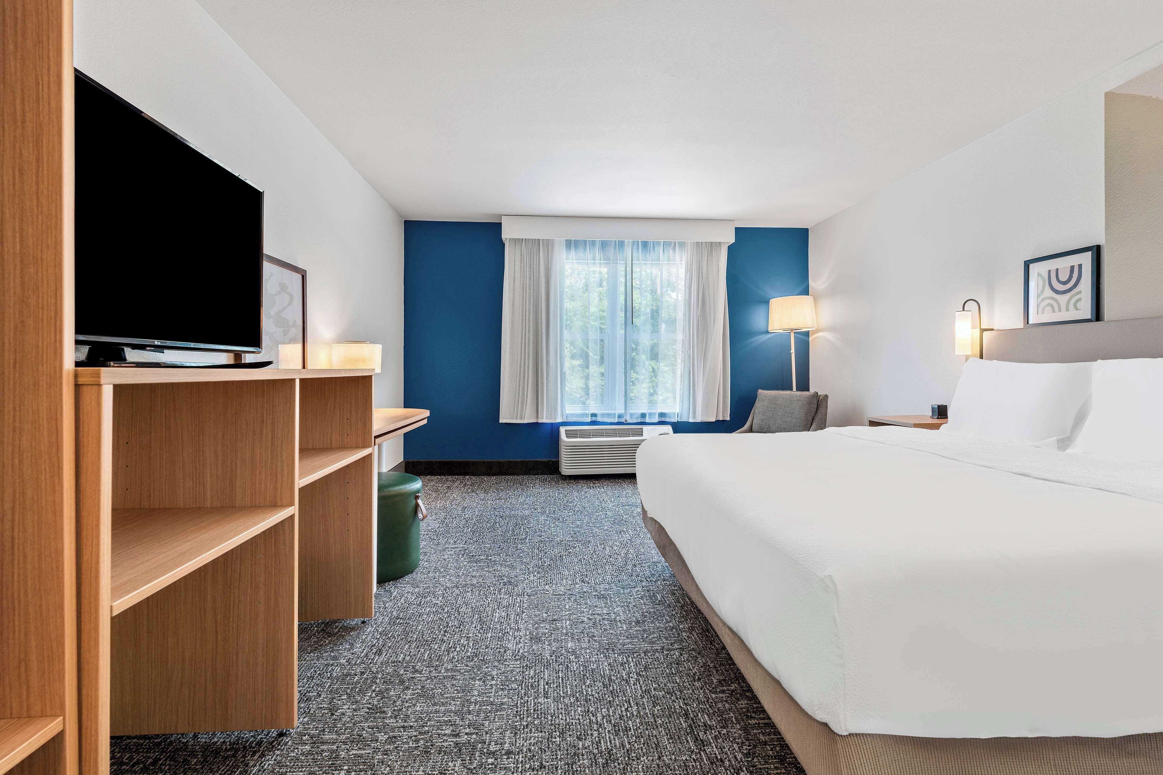 Quarto de hotel com uma cama grande, uma TV de tela plana em uma estante de madeira, uma janela com cortinas brancas, um abajur e uma obra de arte pendurada na parede.