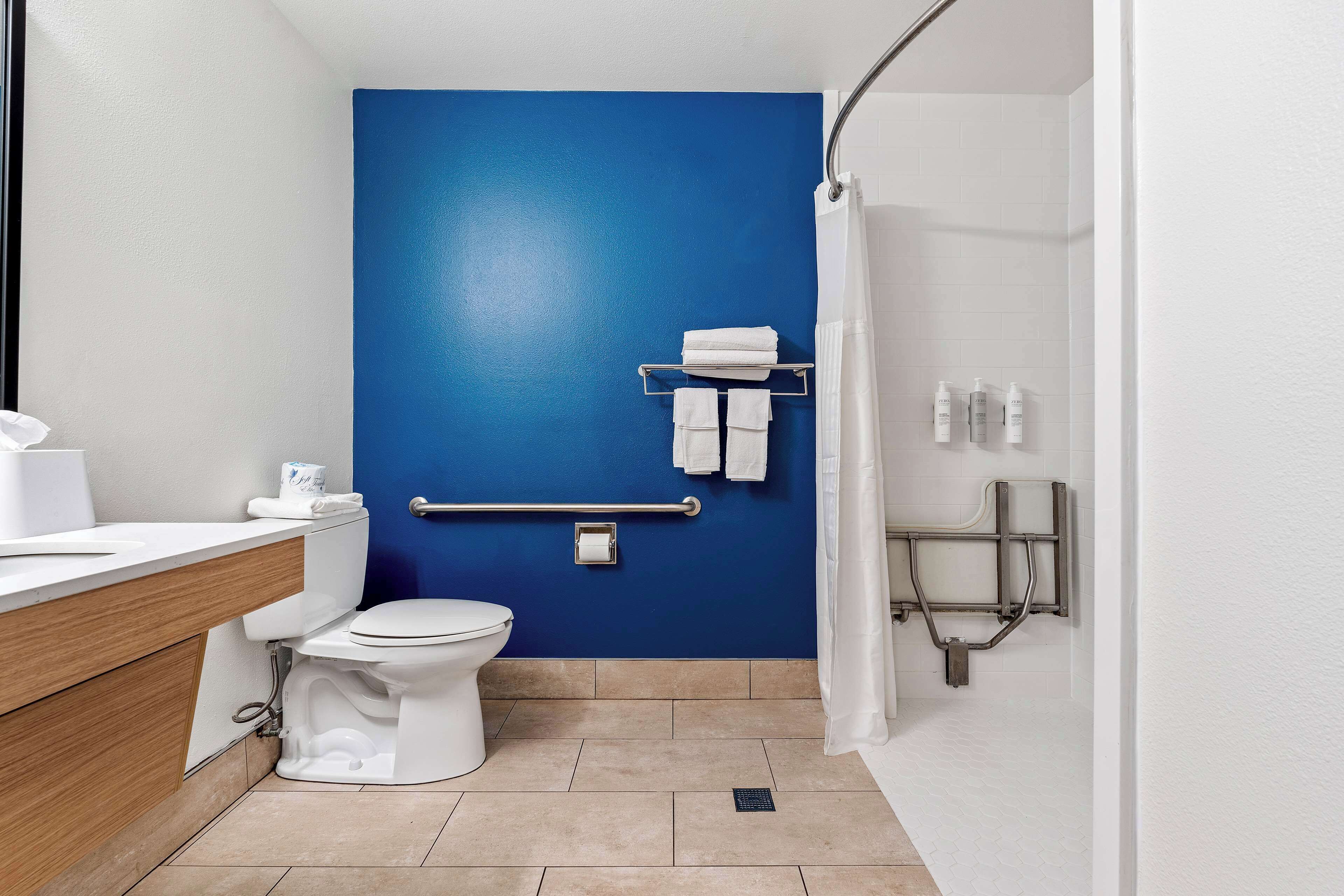 A bathroom with a blue accent wall, a white toilet, a sink with a wooden vanity, a shower area with a grab bar, and a towel rack with towels.