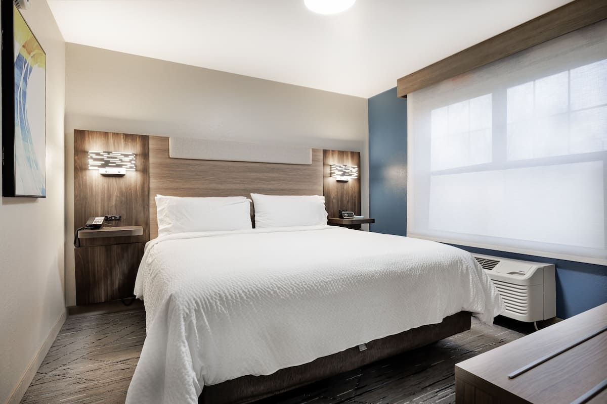 A modern hotel room with a large bed, bedside tables, a wall-mounted phone, and a window with blinds.