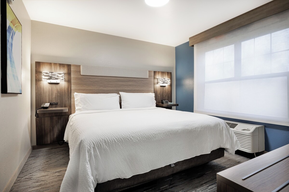 A hotel room with a large bed, bedside tables, a wall-mounted phone, and a window with blinds.
