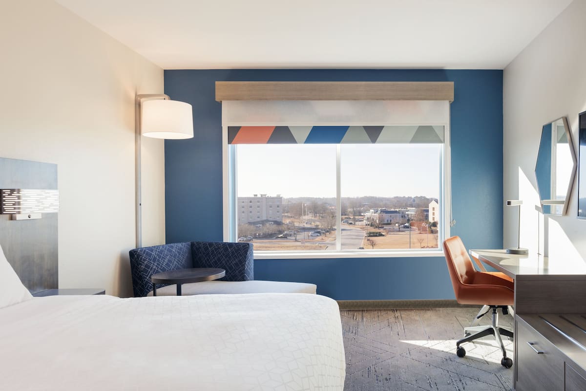 A hotel room with a bed, a desk, a chair, a lamp, and a large window with a view of buildings and a parking lot.