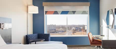 A hotel room with a bed, a desk, a chair, a lamp, and a large window with a view of buildings and a parking lot.