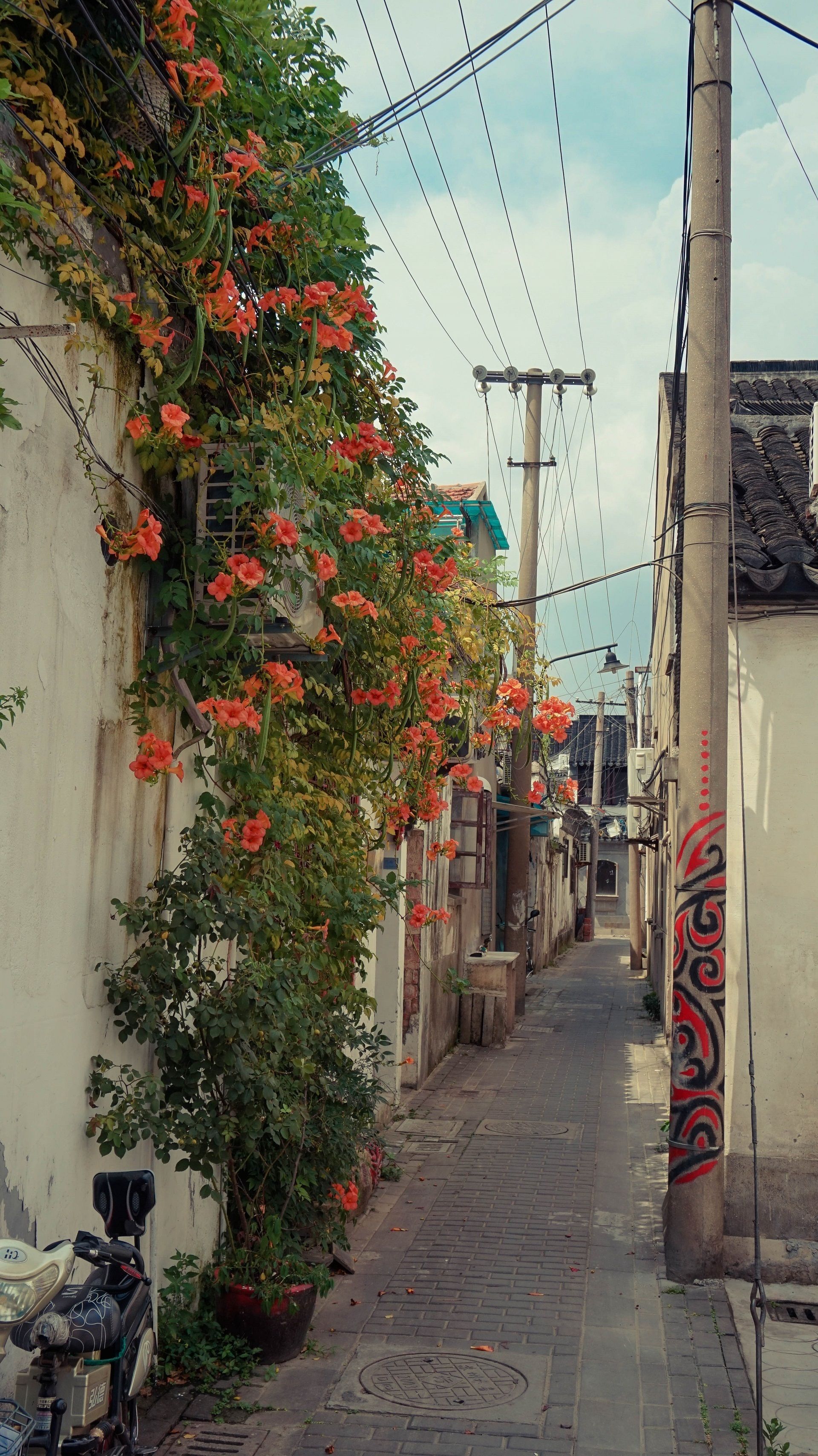 A narrow alleyway with a row of buildings on one side, a parked scooter, and a wall with red graffiti.