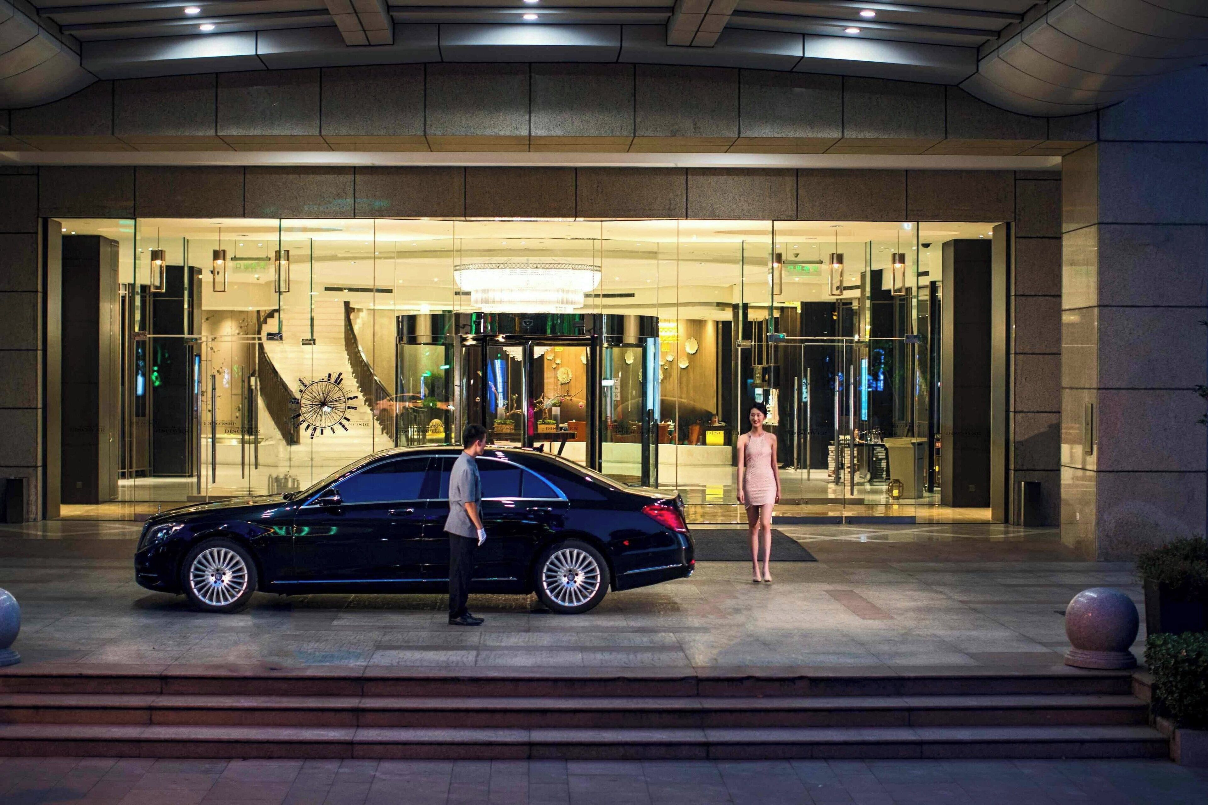 A black car parked in front of a building with a glass entrance, a staircase, and a chandelier.