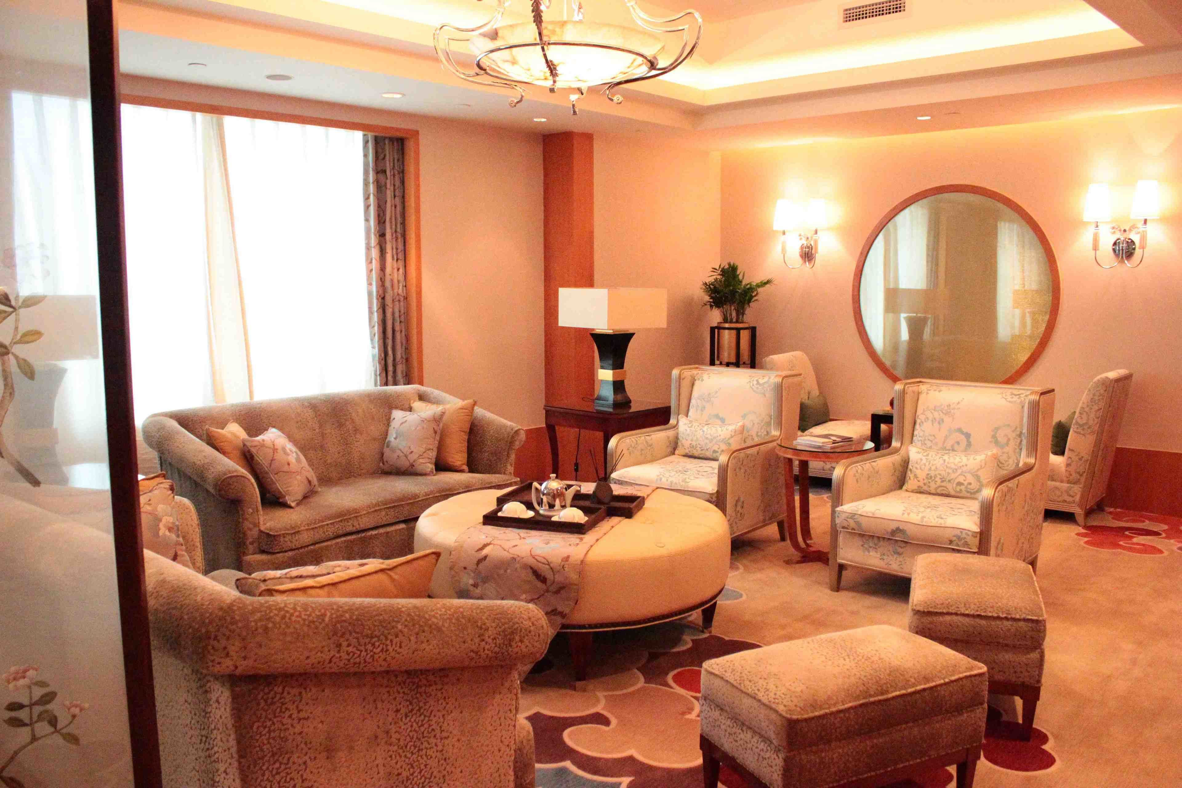 A hotel room with a sofa set, armchairs, a round coffee table, a side table with a lamp, a mirror, and a large window with curtains.