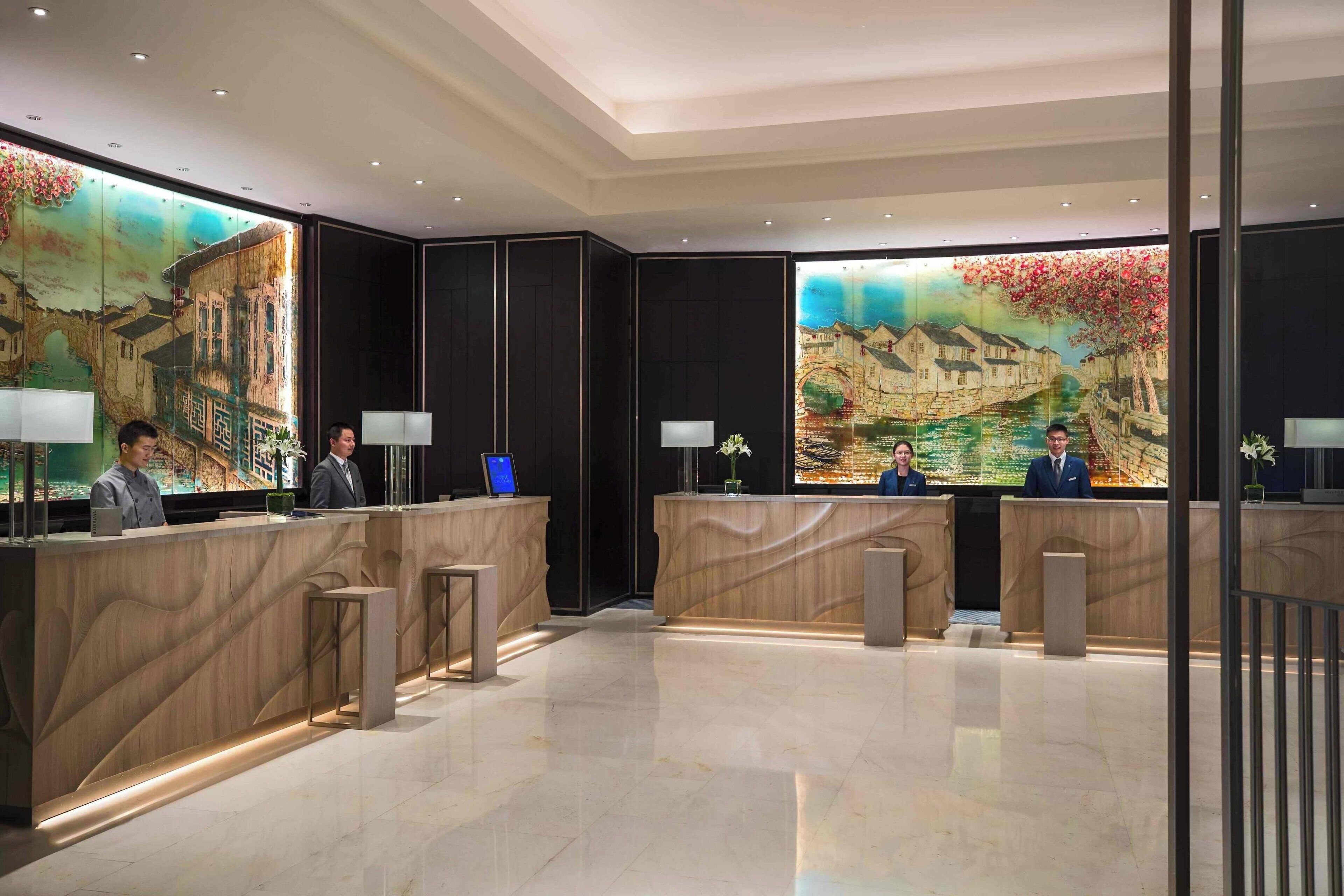 A hotel reception area with two reception desks, each with a receptionist. The reception area features a large mural on the back wall depicting a historical scene.