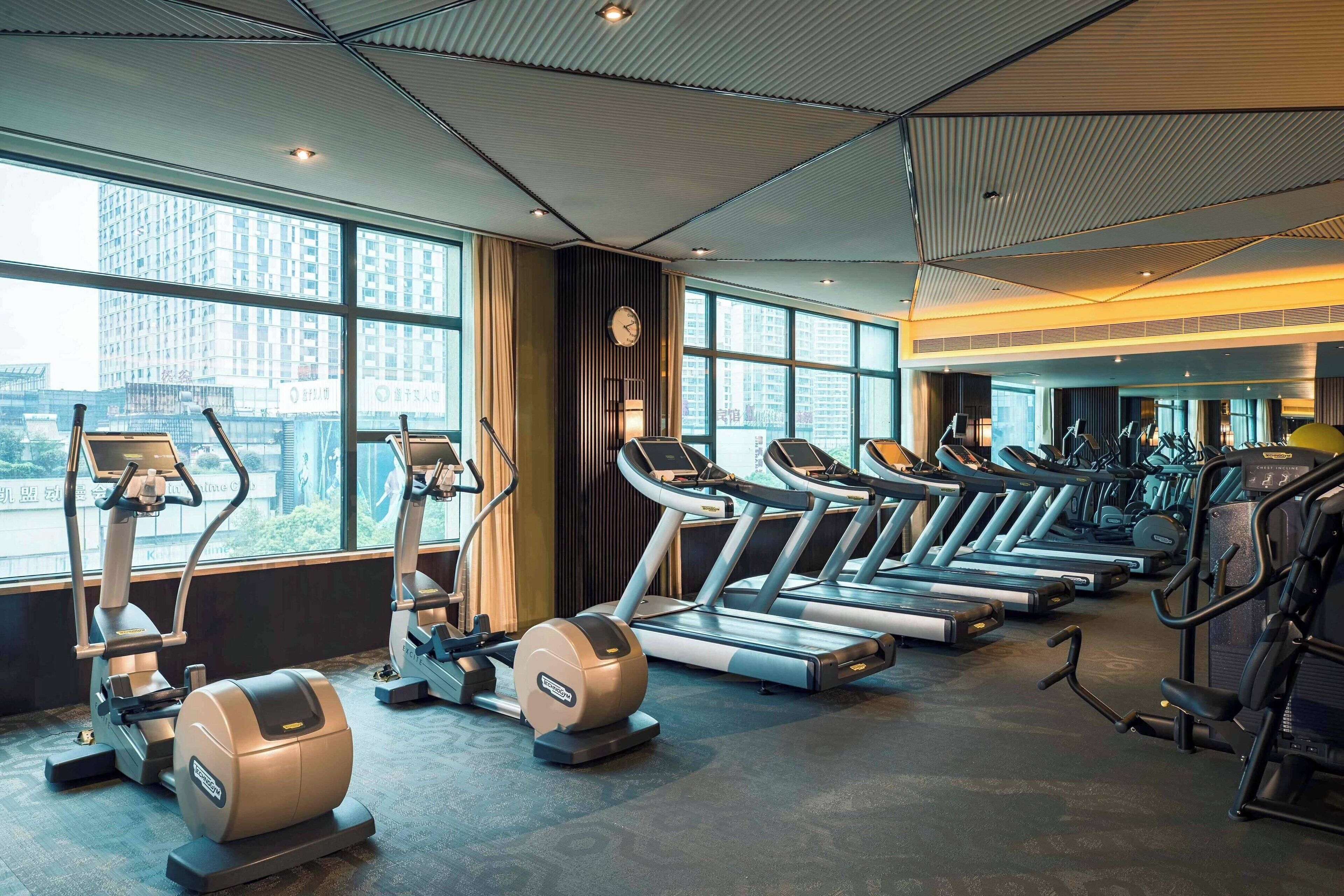 A gym with treadmills, ellipticals, and stationary bikes, large windows, and a modern ceiling design.