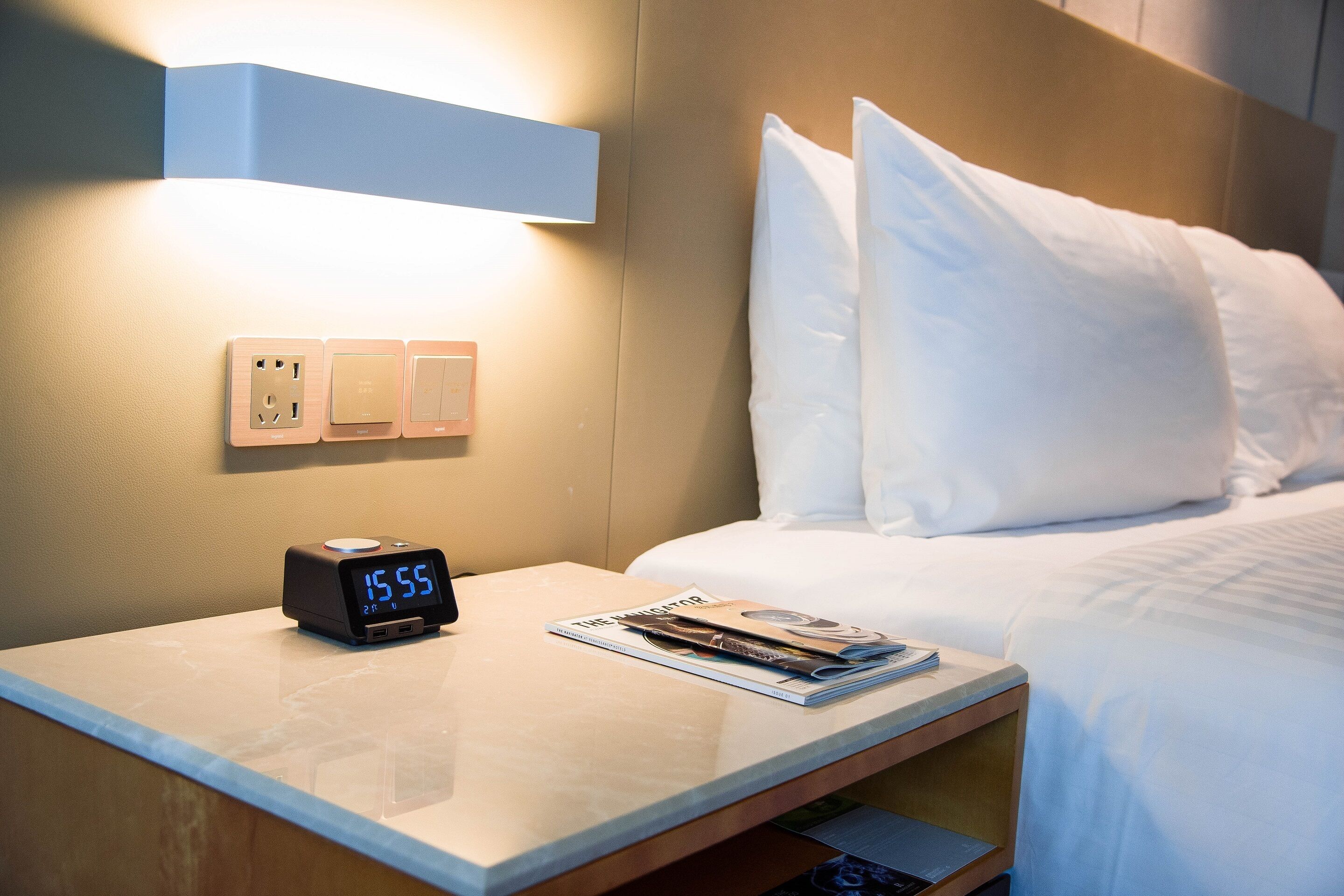 A neatly made bed with white linens, a bedside table with a digital clock, and a magazine.