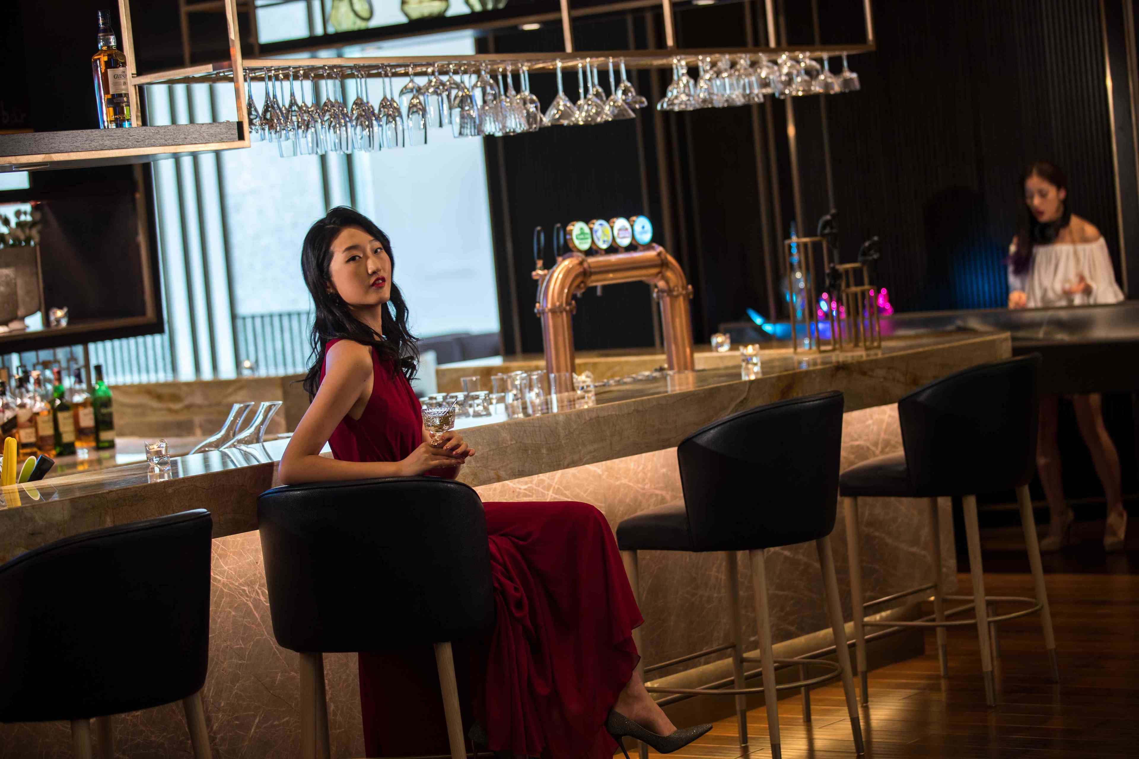 A bar area with a woman in a red dress seated at a bar stool, a beer tap, and a bartender in the background.