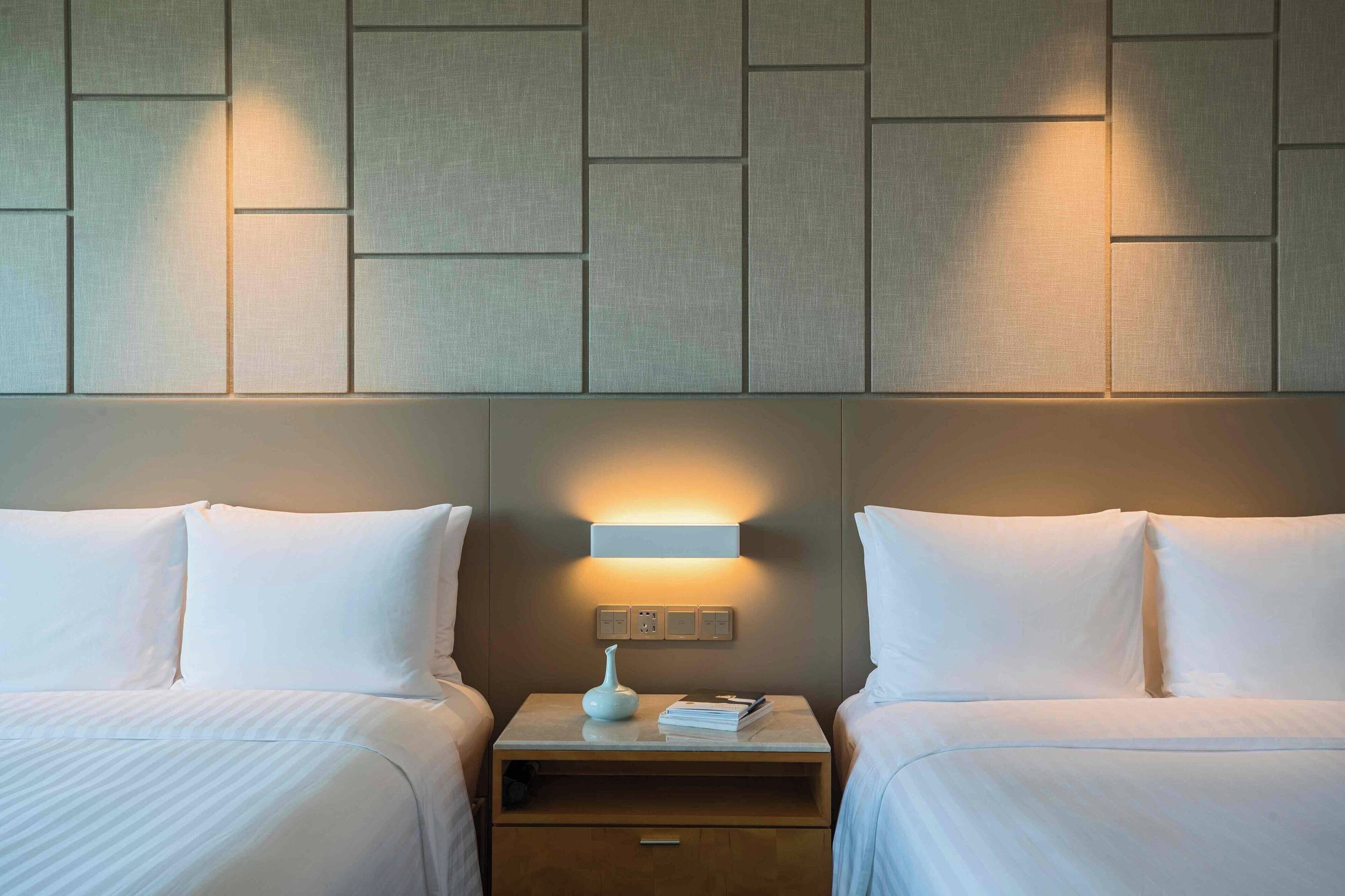 Quarto de hotel com duas camas, uma mesa de cabeceira com abajur e painel decorativo na parede.