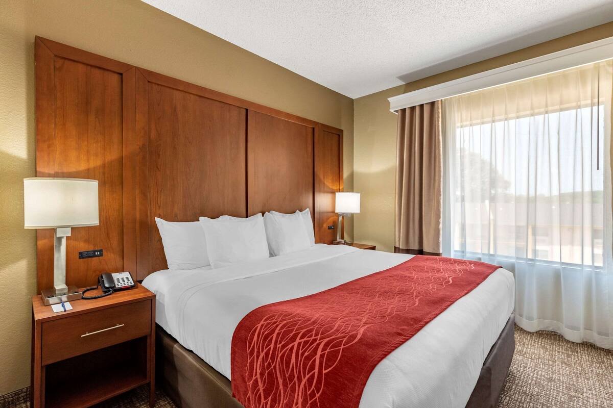 A hotel room with a large bed, wooden headboard, two bedside lamps, a telephone, and a window with sheer curtains.