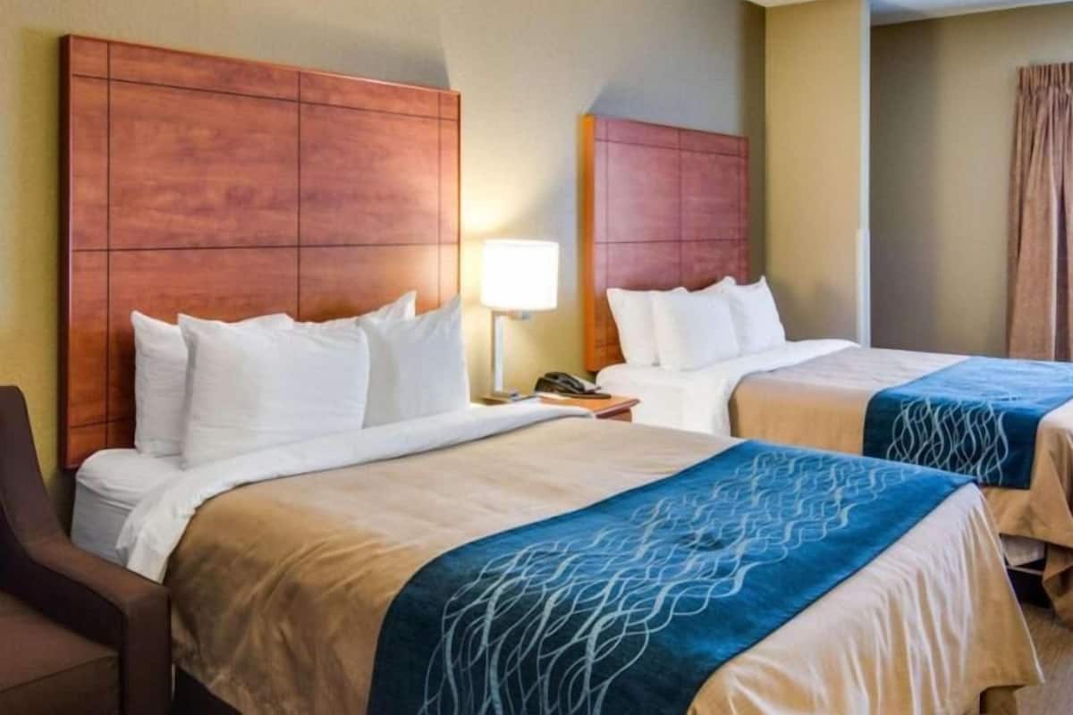 A hotel room with two beds, a desk, a chair, and a lamp.