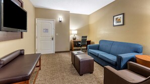 Habitación de hotel con un banco de cuero marrón, un sofá azul, un reposapiés marrón, un televisor de pantalla plana montado en la pared, un escritorio con silla y un cuadro en la pared.