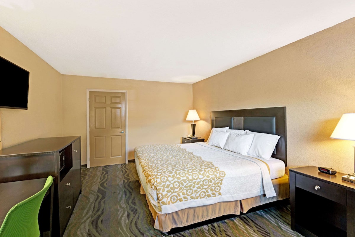 A hotel room with a bed, a desk, a chair, a nightstand with a lamp, and a door.