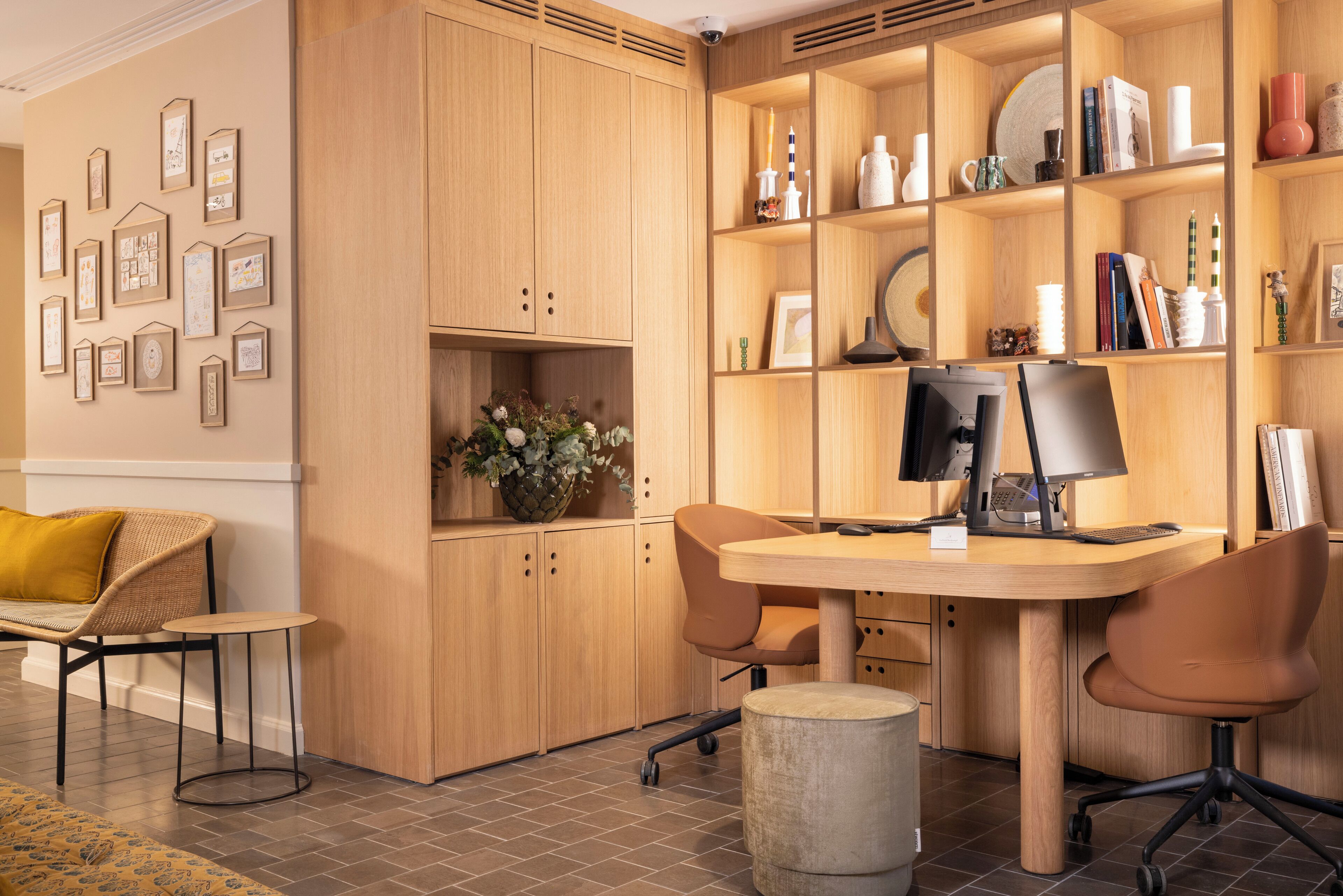 A modern office space with a wooden desk, two chairs, a computer, and a bookshelf filled with books and decorative items.