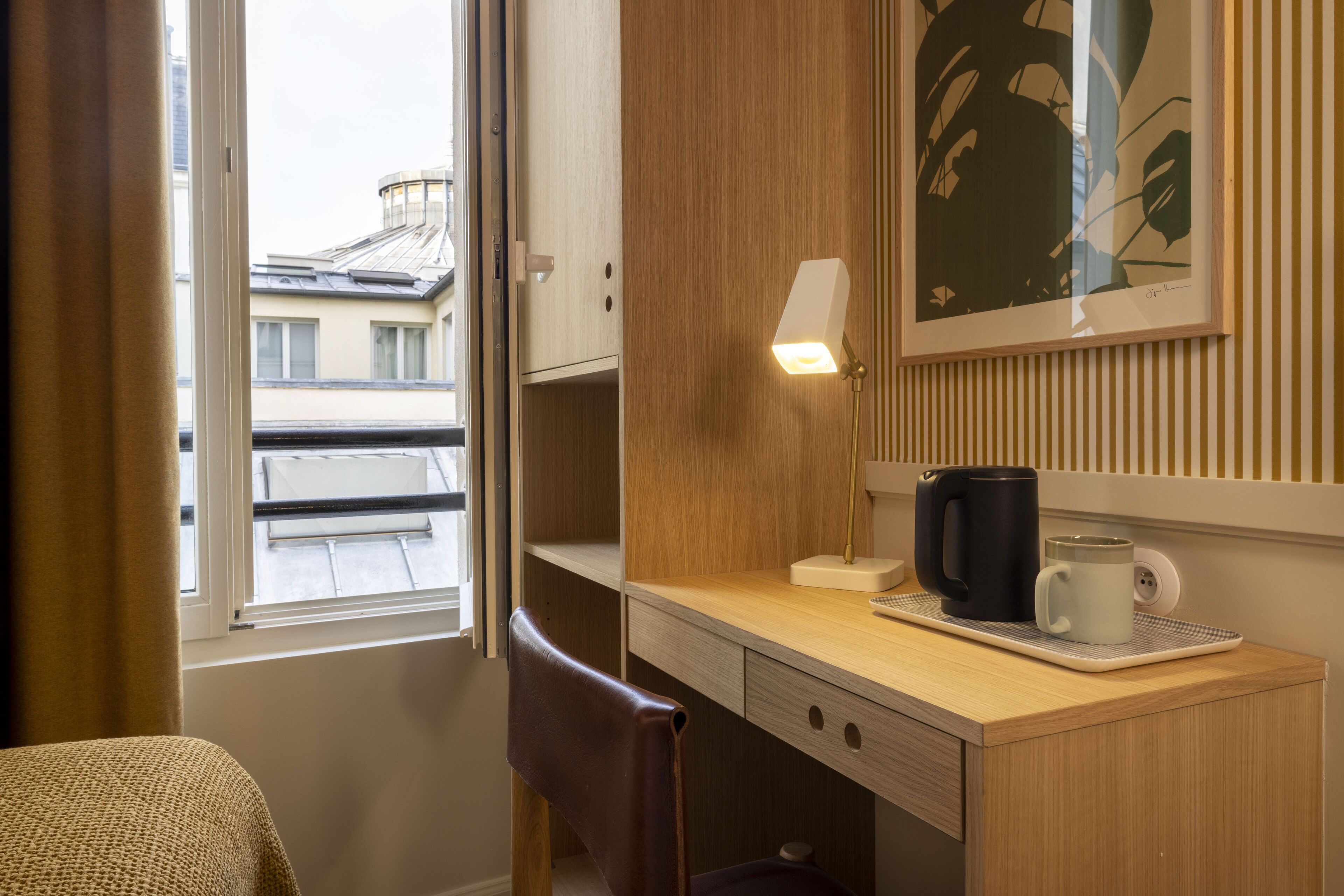 Ein Hotelzimmer mit einem Holztisch, einem Sessel, einem Fenster mit Vorhängen, einem gerahmten Kunstwerk und einer Kaffeemaschine.