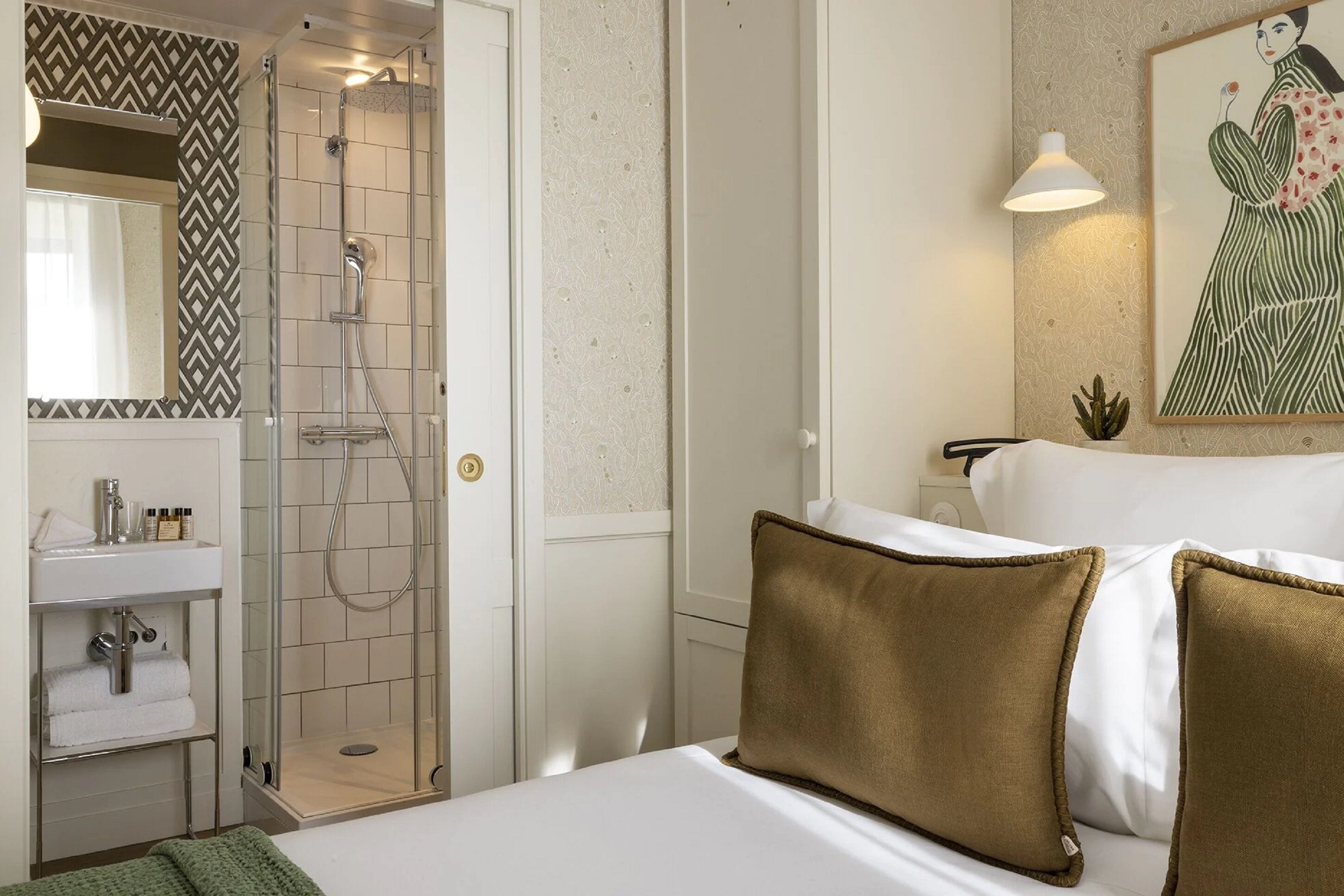 A hotel room with a shower, a sink, a mirror, a bed with pillows, and a painting on the wall.
