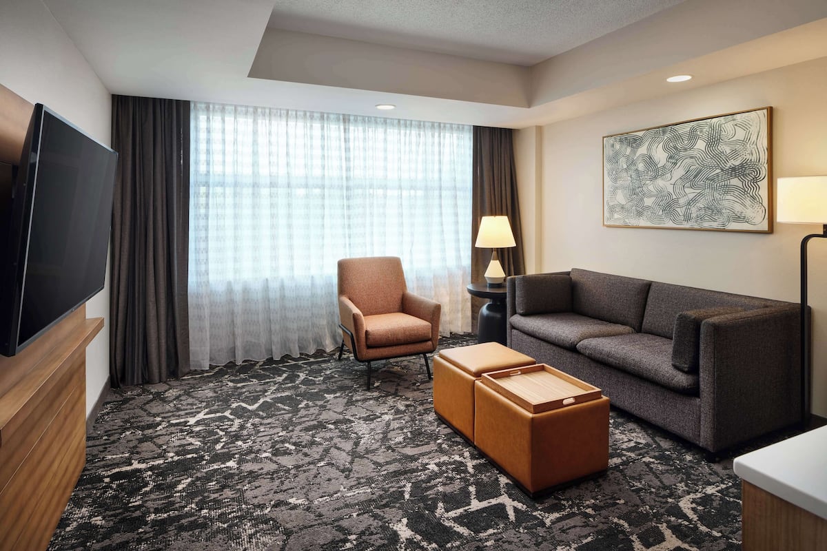 A hotel room with a large flat-screen TV, a sofa, a chair, a coffee table, and a large window with curtains.
