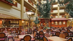 A spacious indoor dining area with multiple tables and chairs, a large central tree, and a kitchen in the background. - Embassy Suites by Hilton Hampton Convention Center (Hampton)