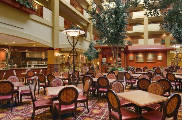 A spacious indoor dining area with multiple tables and chairs, a large central tree, and a kitchen in the background.