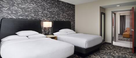 Habitación de hotel con dos camas, un papel tapiz estampado, una mesita de noche con lámpara y vista al pasillo.