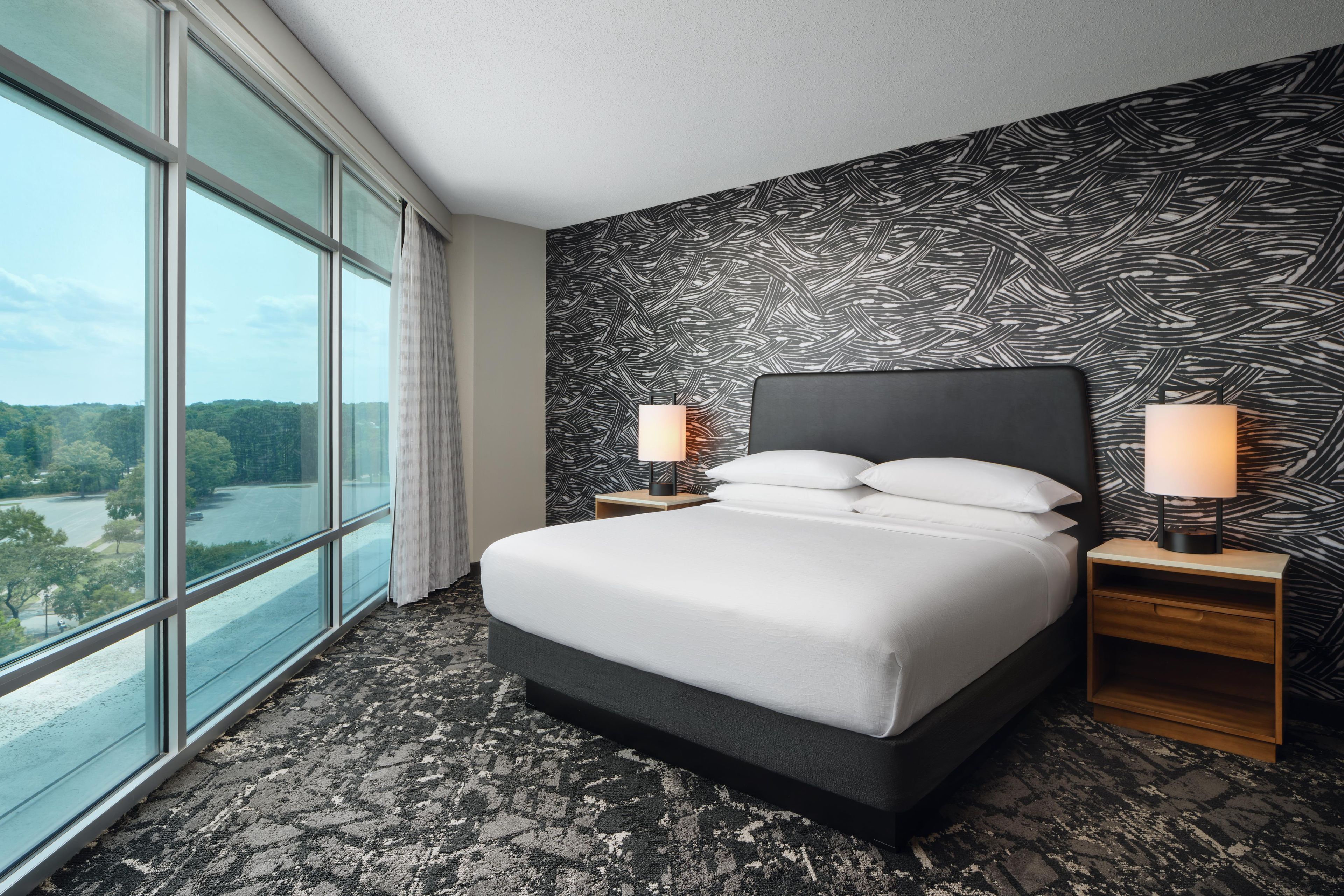 Une chambre d’hôtel avec une grande fenêtre, un lit recouvert de draps blancs, deux tables de chevet avec des lampes et un mur orné d’un motif.