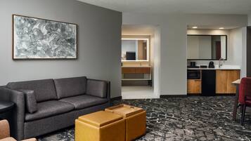 Quarto de hotel com sofá cinza, um banquinho de madeira, tapete estampado em preto e branco, cozinha compacta com micro-ondas e um espelho grande.