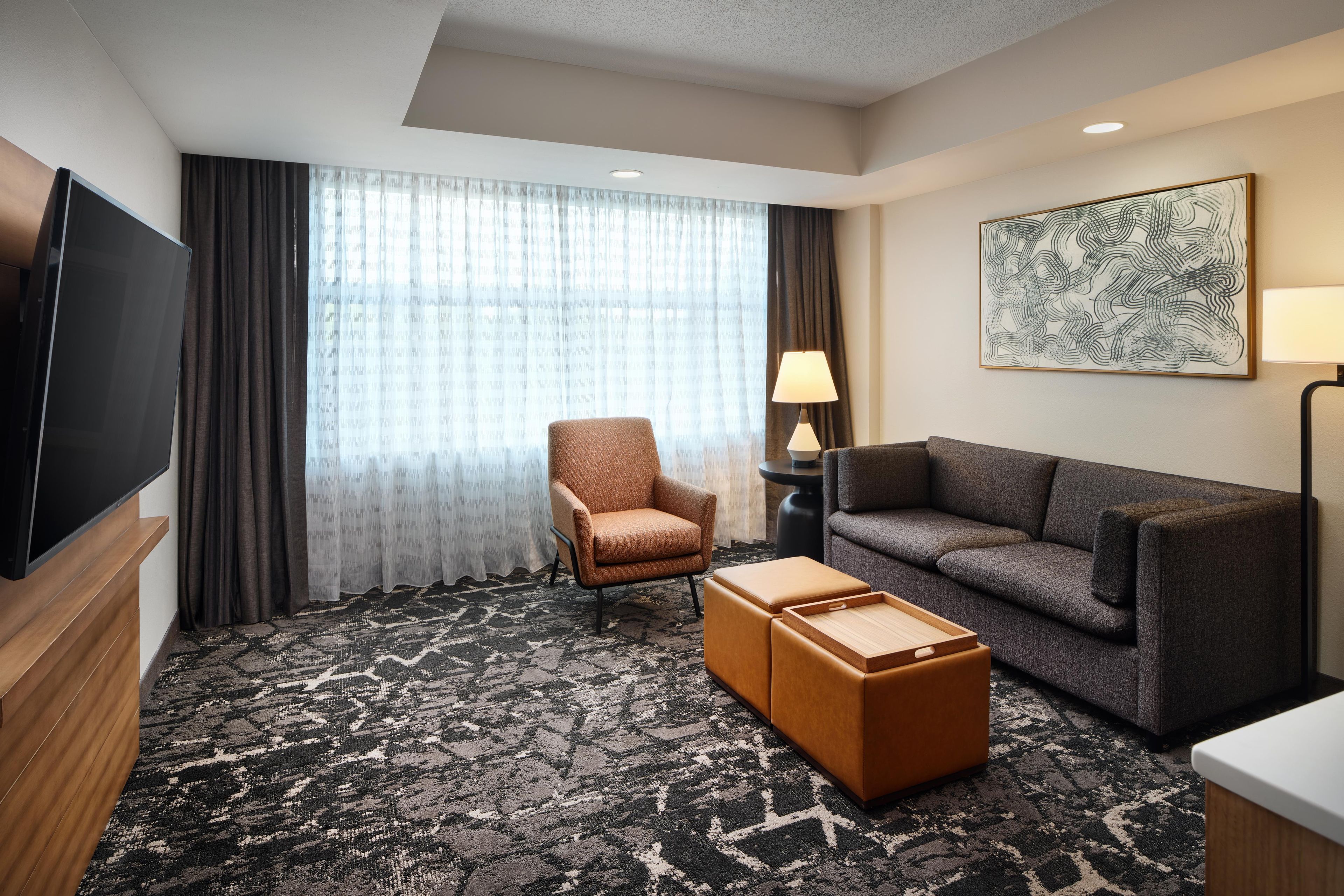 A hotel room with a flat-screen TV, a brown armchair, a grey sofa, a patterned carpet, and a painting on the wall.
